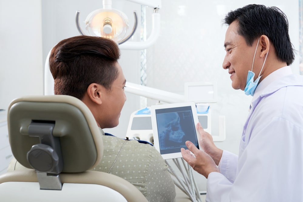 asian-male-dentist-demonstrating-jaw-x-ray-image-tablet-patient (1)-min